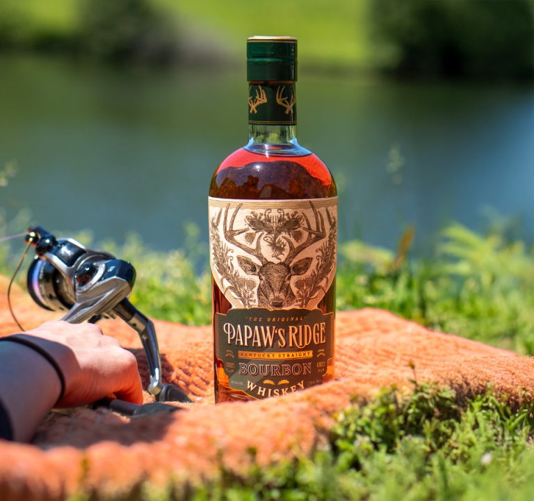 Bottle of PaPaw's Ridge bourbon resting on a blanket with a hand resting on a fishing pole next to it, in front of a small pond