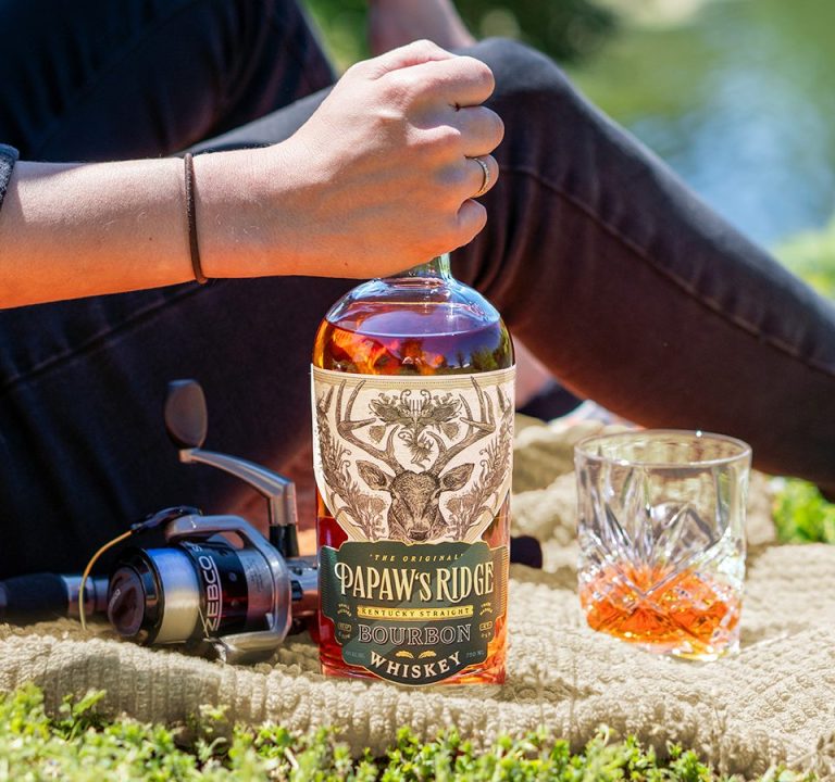 Photo of PaPaw's Ridge Bourbon being held by a woman sitting down on a blanket with a glass of bourbon and fishing pole in front of a pond.