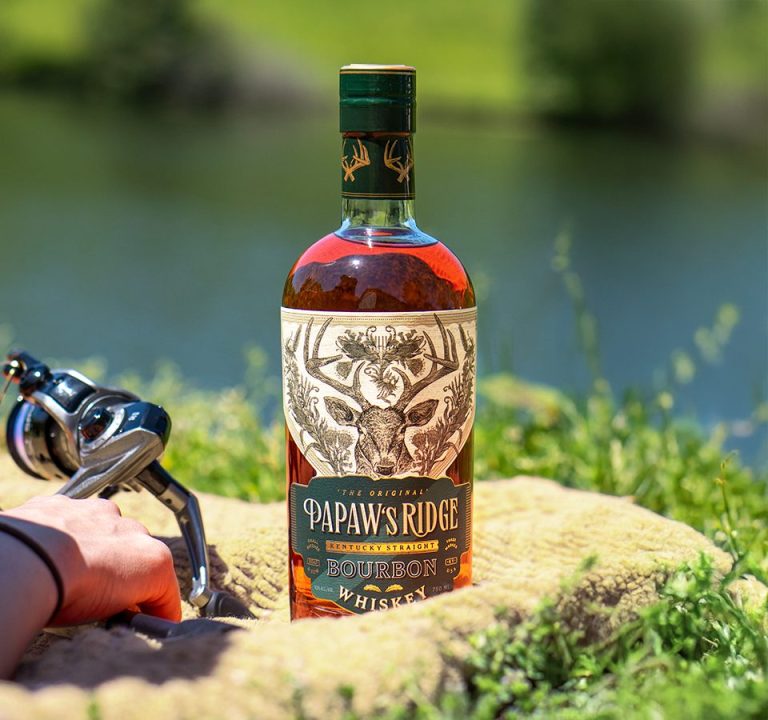 Photo of PaPaw's Ridge Bourbon sitting on blanket in front of pond with hand resting on a fishing pole in the left of the frame.